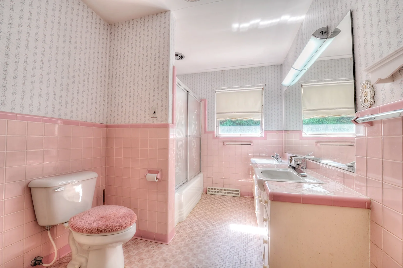 Dated 1950s bathroom with pink wall tile, pink floral wallpaper, a bathtub behind sliding glass doors, and a pink toilet